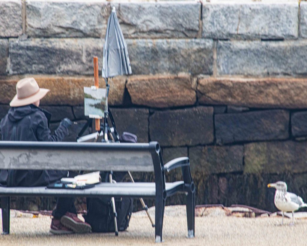An artist painting outdoors in Rockport, Massachusetts, with a seagull standing nearby as an observer