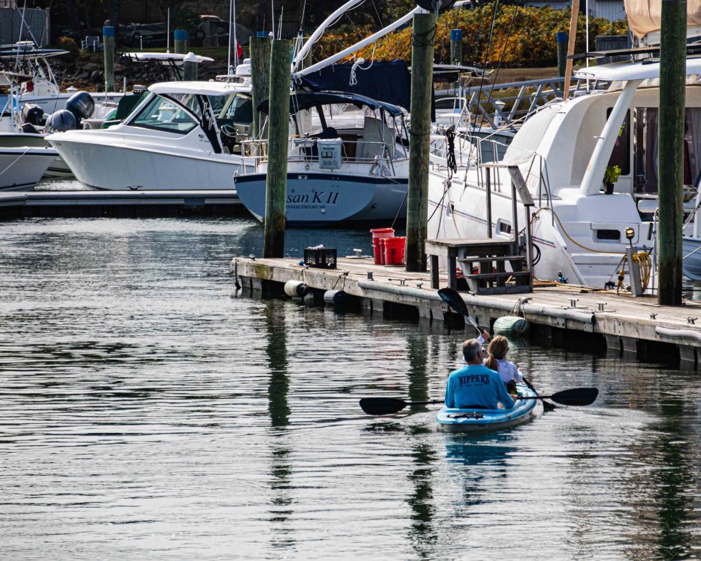 Two people kayaking through Kennebunkport Harbor, passing by docked boats.