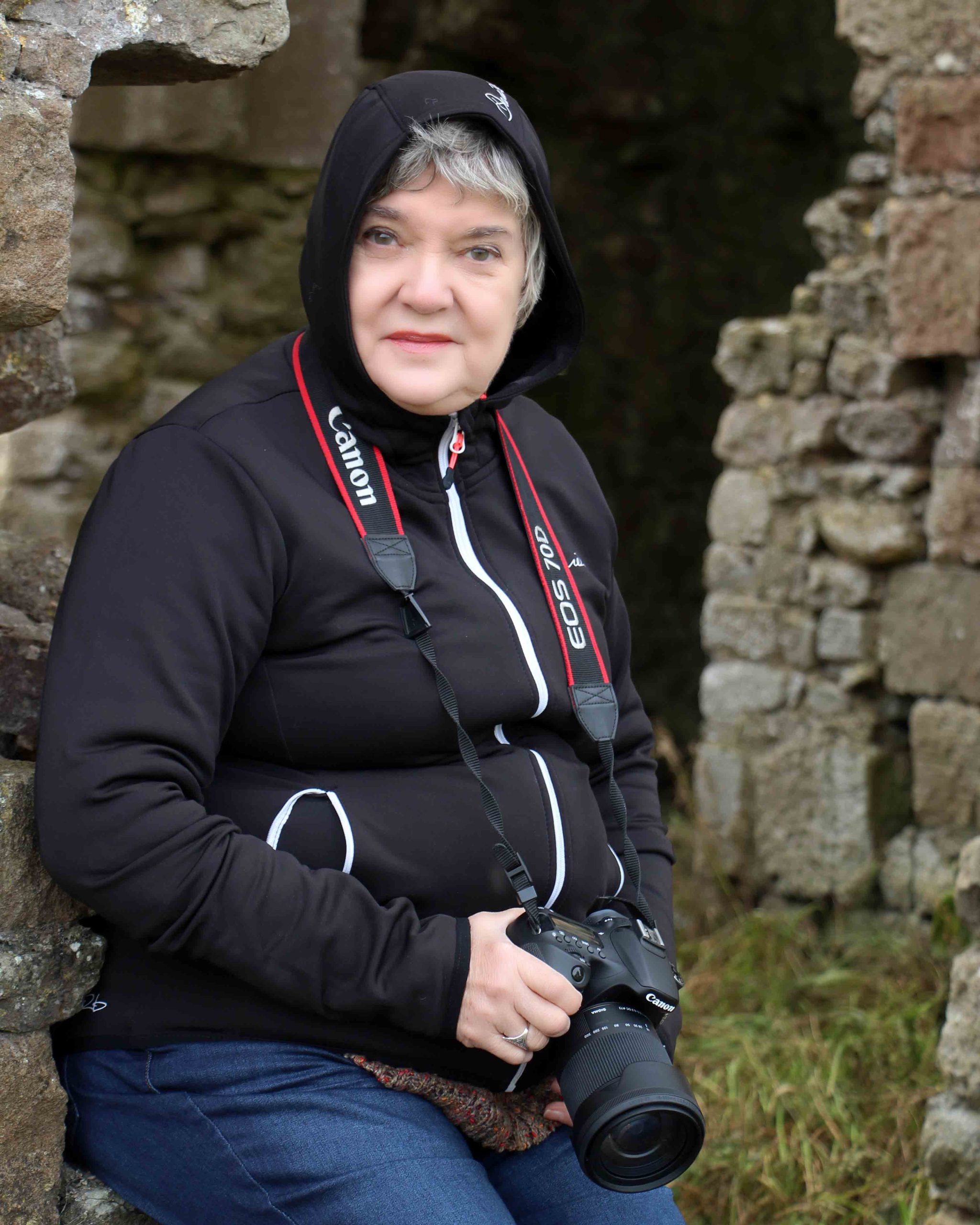 Portrait of Sandy Hitz holding a camera, sitting by stone ruins, highlighting her passion for photography.