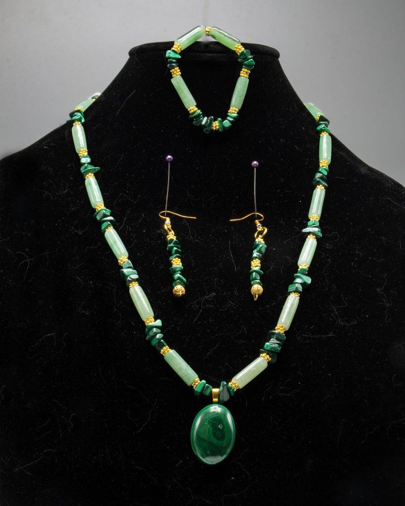 A handcrafted jewelry set featuring a green malachite necklace, bracelet, and matching earrings with gold accents displayed on a black velvet stand.